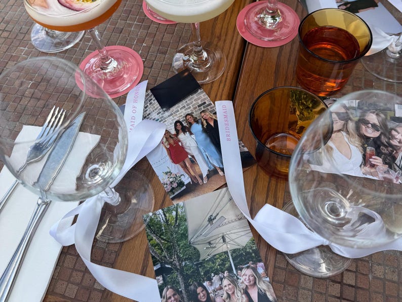 May include: A table setting with cocktails, glassware, and photos. Clear wine glasses with white ribbons, a fork and knife, and amber-colored glasses are visible. Pink coasters and photos with the text "Maid of Honor" are also present.