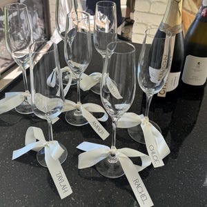 May include: Champagne flutes with white ribbons and name tags, including "Taylah", "Amber", "Michelle", "Chloe", and "Rachel". Two bottles of champagne are in the background. The flutes are arranged on a black surface.