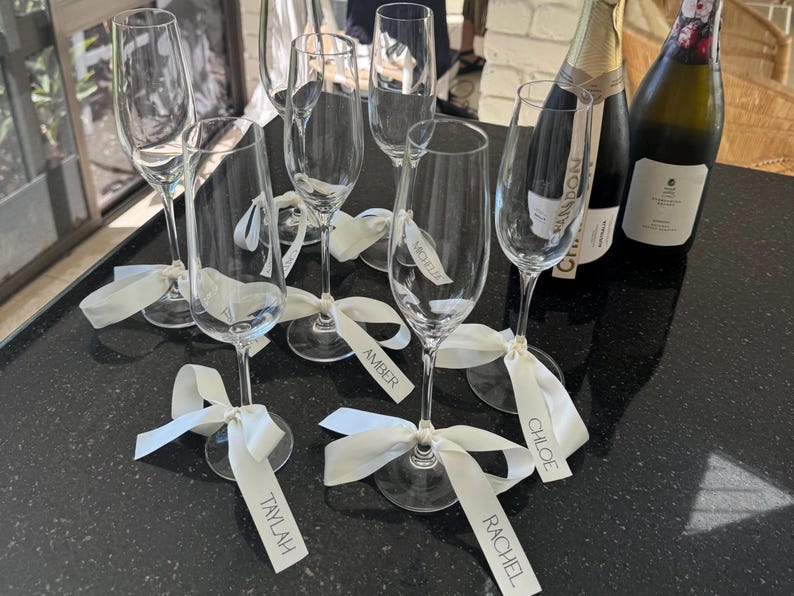 May include: Champagne flutes with white ribbons and name tags, including "Taylah", "Amber", "Michelle", "Chloe", and "Rachel". Two bottles of champagne are in the background. The flutes are arranged on a black surface.
