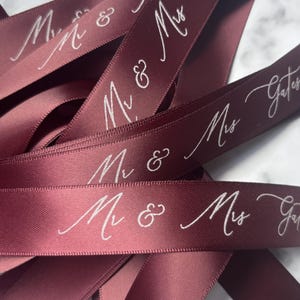 May include: Burgundy satin ribbon with white script text reading "Mr & Mrs Gates". The ribbon is folded and layered, creating a textured effect. Ideal for wedding favors, gift wrapping, or crafting projects.