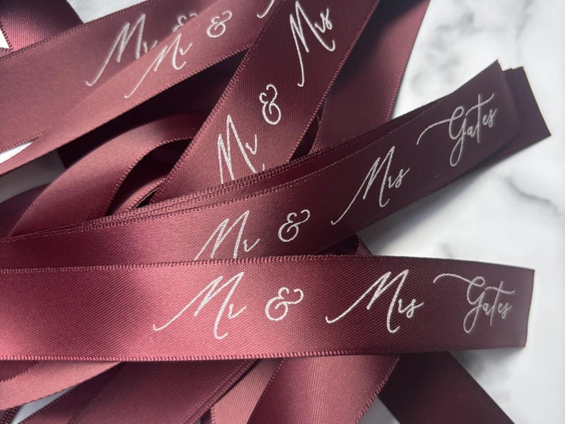 May include: Burgundy satin ribbon with white script text reading "Mr & Mrs Gates". The ribbon is folded and layered, creating a textured effect. Ideal for wedding favors, gift wrapping, or crafting projects.