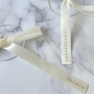 May include: Ivory satin ribbons with gold lettering reading "CHRISTIANNE" and "INGRID" are tied to the stems of clear wine glasses. The ribbons are arranged on a white and gray marble surface.