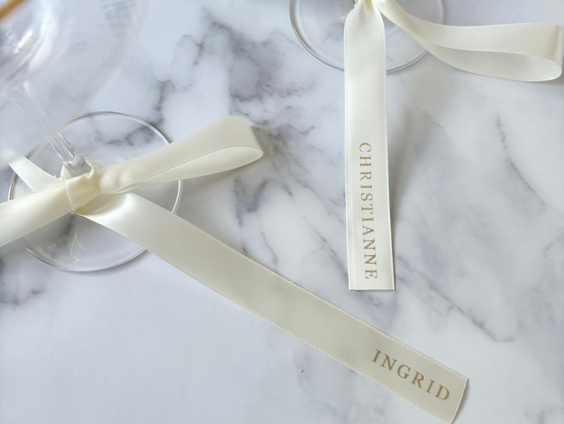 May include: Ivory satin ribbons with gold lettering reading "CHRISTIANNE" and "INGRID" are tied to the stems of clear wine glasses. The ribbons are arranged on a white and gray marble surface.