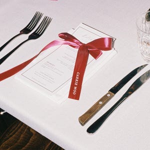 May include: A table setting with two black forks, two knives, a water glass, and a menu tied with a red ribbon. The ribbon has the text "CARRIE WOO" printed on it. The menu is white with black text.