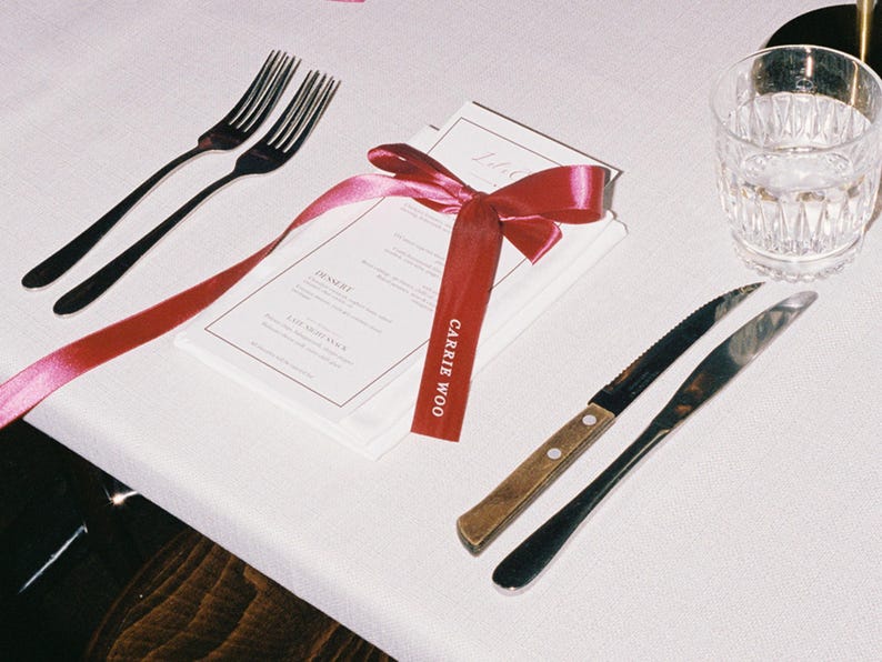 May include: A table setting with two black forks, two knives, a water glass, and a menu tied with a red ribbon. The ribbon has the text "CARRIE WOO" printed on it. The menu is white with black text.