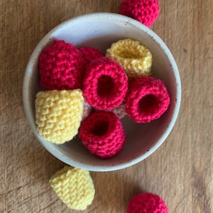 May include: A white bowl filled with crocheted raspberries and lemons. The raspberries are red and the lemons are yellow. There are also a few crocheted raspberries and lemons outside of the bowl.