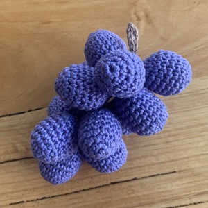 May include: A crocheted bunch of grapes in a purple shade. The grapes are connected to a stem made of a light grey yarn.