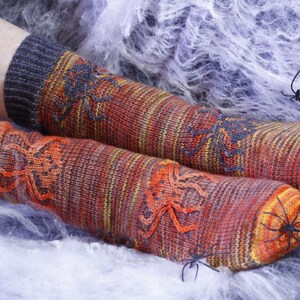 Puede incluir: Un par de calcetines a rayas naranja, amarillo y gris con diseños de arañas negras. Los calcetines están sobre un fondo de telaraña blanca.