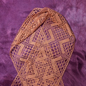 May include: A delicate, crocheted shawl in a warm brown colour, draped on a purple fabric background. The intricate lace pattern features a repeating floral motif.