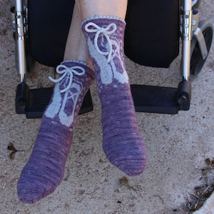 Puede incluir: Un par de calcetines de punto morado y blanco con un diseño de zapato de ballet. Los calcetines tienen lazos blancos en la parte superior.