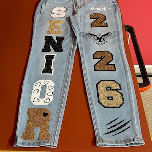 Pode incluir: Jeans azul claro com letras e gráficos decorativos. A perna esquerda apresenta a palavra "SENIOR" em uma mistura de letras brancas, pretas e douradas com glitter. A perna direita tem "class of 2026" e um gráfico de águia.