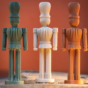 May include: Three wooden figurines in green, cream, and brown tones are displayed against an orange backdrop. Each figure features a cylindrical head, torso, arms, and legs, all with vertical grooves. The figurines are presented on a wooden surface.