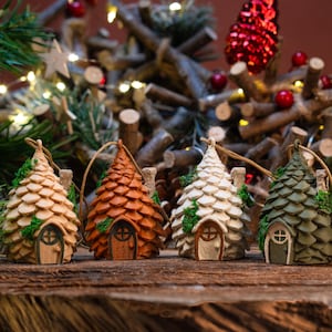 Op de afbeelding: Vier miniatuur kerstboomornamenten op een rij, elk met een unieke kleur: beige, oranje, wit en groen. Elk ornament heeft een kleine deur en een schoorsteen, met een rustieke, sprookjesachtige esthetiek. De achtergrond toont een kerstboom en lichtjes.