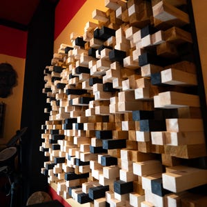 Puede incluir: Panel acústico de pared hecho de bloques alternos de madera natural y pintados de negro. Los bloques están dispuestos en un patrón escalonado, creando una superficie texturizada. La pared está montada en una pared amarilla con molduras rojas.