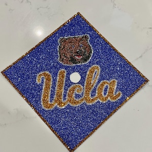 May include: A blue graduation cap with glitter, featuring the word "Ucla" in gold script and a brown bear illustration. The cap has a gold border and a white button in the center.