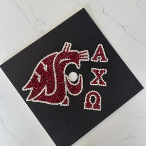 May include: Black graduation cap decorated with a red and silver glitter design. The design includes a stylized cougar logo and the Greek letters A, X, and Ω, all outlined in silver rhinestones.