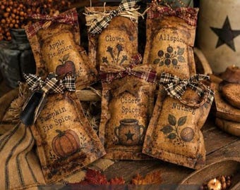 May include: A collection of six rectangular fabric sachets with a distressed tan finish, each tied with a plaid ribbon. The sachets are labeled Apple Pie Spice, Cloves, Allspice, Pumpkin Pie Spice, Cider Spice, and Nutmeg, featuring printed illustrations of the corresponding ingredients.