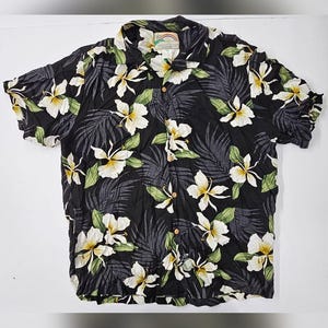 May include: A black short-sleeve button-down shirt featuring a repeating pattern of white plumeria flowers with yellow centers and dark gray palm fronds. The shirt has a collar and a label at the neckline that reads Paradise Found with a rainbow.