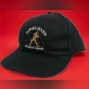 May include: A black baseball cap featuring an embroidered logo on the front. The logo includes the text HORMEL FOODS in white, a gold-coloured graphic of a baseball player swinging a bat, and the text Baseball Classic in white lettering.