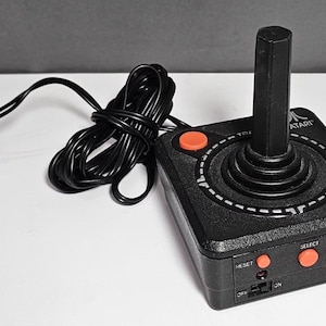 Puede incluir: Un mando de control Atari negro con una palanca central vertical, un botón de acción naranja en la parte superior y tres botones naranjas más pequeños etiquetados como Reset, Select y Start en el borde frontal con un interruptor de encendido.