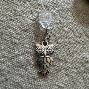 May include: A silver-toned metal owl charm suspended from a clear plastic clip. The owl features detailed engraved wings, a pointed beak, and large circular eyes with radial lines. The charm is attached to the clip by a small silver metal jump ring.