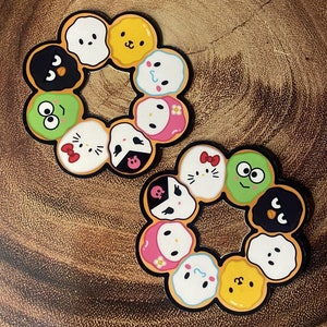 May include: Two donut-shaped stickers with different cartoon characters. Each sticker has a yellow, white, black, pink, green, and light blue character. The characters are arranged in a circle.