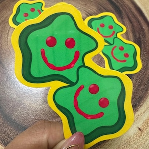 May include: A collection of green, star-shaped stickers with yellow and dark green borders. Each sticker features a red smiling face with two red dots above the smile. The stickers are held in a hand.