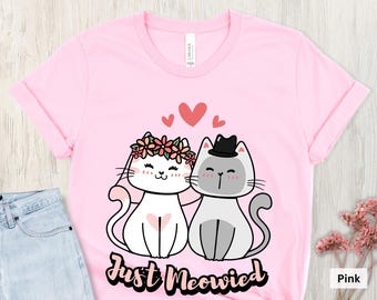 May include: Pink t-shirt featuring a cartoon illustration of two cats, one wearing a floral crown and the other a hat, with hearts above them. The text "Just Meowied" is below the cats. The shirt is pink.