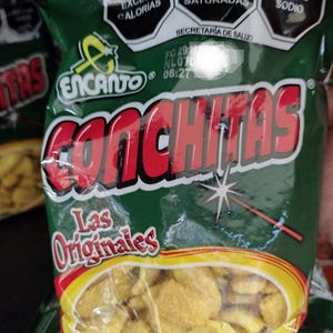 May include: A green bag of "Encanto Conchitas" tortilla chips. The bag has red and yellow accents, with text in Spanish indicating high calorie, saturated fat, and sodium content. The chips are visible through the clear plastic.