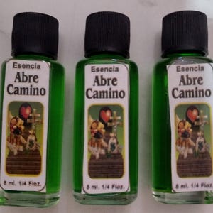 May include: Three small green glass bottles with black caps. Each bottle has a white label with the words "Esencia Abre Camino" and an illustration of a heart and cross. The bottles contain 8 ml of liquid.