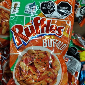 May include: A bag of Ruffles potato chips with a salsa buffalo flavor. The bag is orange and green with the Ruffles logo and an image of the chips. The bag also has the Mexican flag and FIFA logo.