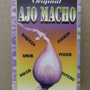 May include: A rectangular box of "JABON Original AJO MACHO" soap. The box features a purple, orange, and yellow gradient background with a purple onion graphic. Text includes "SALUD", "AMOR", "RIQUEZA", "FUERZA", "PODER", and "DINERO".