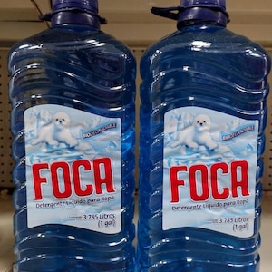 May include: Two large, clear plastic jugs filled with blue liquid laundry detergent. Each jug features a label with a white seal, the text FOCA, Detergente Líquido para Ropa, and a net content of 3.785 Litros (1 gal) with a blue screw-on cap.