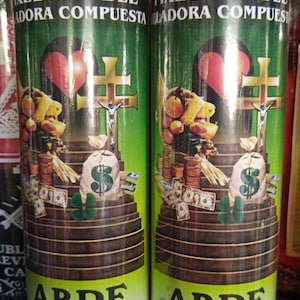 May include: Two tall cylindrical glass candles with green labels featuring the text FIXED CANDLE, VELADORA COMPUESTA, and ABRE CAMINO. The labels display a graphic of a cross, a heart, a sack with a dollar sign, stacks of coins, and paper money.