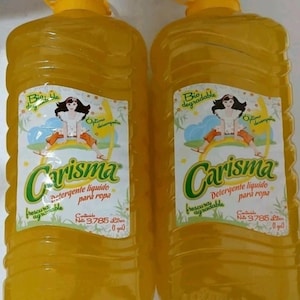 May include: Two large, yellow plastic bottles of laundry detergent. Each bottle has a yellow cap and a label with the word "Carisma" in green, along with an illustration of a woman and text in Spanish. Each bottle contains 3.785 liters (1 gal).