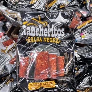 May include: A black bag of Rancheritos Salsa Negra tortilla chips. The bag has the brand name in white and yellow, with the words "Salsa Negra" below. The chips are visible through the clear plastic, and the bag has nutritional information.