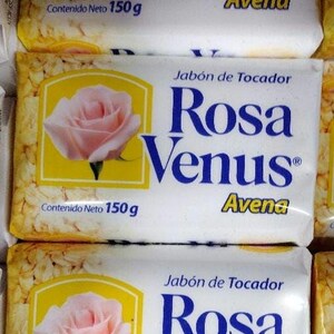 May include: White soap bars with the text "Rosa Venus" in blue, featuring a pink rose illustration. The bars are labeled "Avena" and "Jabón de Tocador". Each bar contains 150g of product.