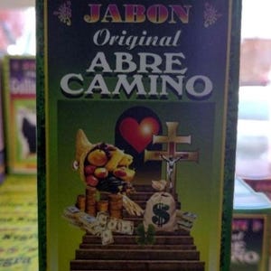 May include: A green and gold box with the words "JABON ORIGINAL ABRE CAMINO" in white and gold lettering. The image features a cornucopia, a red heart, a cross, a money bag, and stacks of money.