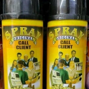 May include: Two spray bottles with black caps and yellow labels. The labels feature the text "SPRAY ORIGINAL CALL CLIENT" in a gold font, along with an image of people and money. The bottom of the label reads "Llama Cliente".