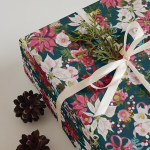 May include: A rectangular gift box wrapped in dark teal paper featuring a floral pattern of pink and white poinsettias, holly berries, and pine needles. A cream-colored ribbon is tied in a bow, accented with a small sprig of green cedar.