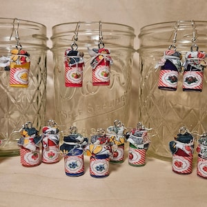 Peut inclure: Une collection de 10 paires de boucles d'oreilles faites à la main avec des bocaux en verre miniatures recouverts de tissu. Les bocaux sont décorés de motifs et de couleurs variés, notamment le rouge, le bleu, le jaune et le vert. Les boucles d'oreilles sont présentées sur une surface en bois.