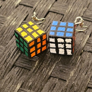 May include: A pair of earrings featuring miniature Rubik's Cube designs. One cube is painted in yellow, orange, green, and black, while the other is painted in blue, white, and black.