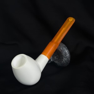May include: A white and orange tobacco pipe with a white bowl and an orange stem. The pipe is resting on a black surface.