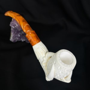 May include: A white meerschaum pipe with intricate carvings and a brown amber mouthpiece. The pipe is shaped like a hand holding a bowl.