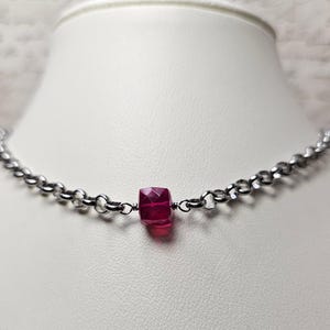 以下が含まれることがあります： ファセット加工が施された深紅の宝石ペンダントが付いた、シルバーカラーのチェーンネックレス。ネックレスは白いマネキンに飾られています。宝石は鮮やかで人目を引く焦点です。