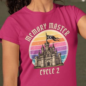 Op de afbeelding: Magenta T-shirt met een retro design. Het ontwerp bevat de woorden "MEMORY MASTER" boven een kasteelillustratie, tegen een gestreepte zonsondergang. De tekst "CYCLE 2" staat eronder.