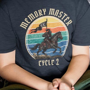 May include: Navy blue t-shirt with a circular graphic featuring a knight on horseback, a flag, and a sunset design. The text "MEMORY MASTER" arches above the image, with "CYCLE 2" below.