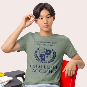 May include: Sage green t-shirt with a navy blue crest design. The crest includes a book, the letter A, the Greek letters Omega and a torch, and the words "CHALLENGE ACCEPTED". The shirt also has the text "FOUNDATIONS LAID" and "ESSENTIALS ADDED".