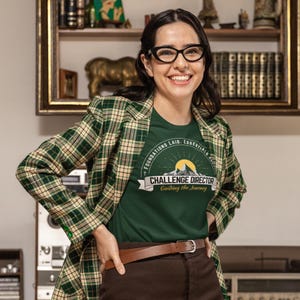 May include: A woman wearing a green t-shirt with the text "CHALLENGE DIRECTOR Guiding the Journey" and a green plaid blazer. The t-shirt features a mountain and sun graphic. She is also wearing brown pants and a brown belt.