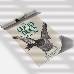 Puede incluir: Un p&oacute;ster enrollado con una detallada ilustraci&oacute;n en blanco y negro de un pato en vuelo. El p&oacute;ster tiene el texto "LUCKY DUCK hunt club" en verde azulado, con "est. 1956" arriba y "home of the single shot, BRITISH COLUMBIA, CANADA" abajo.